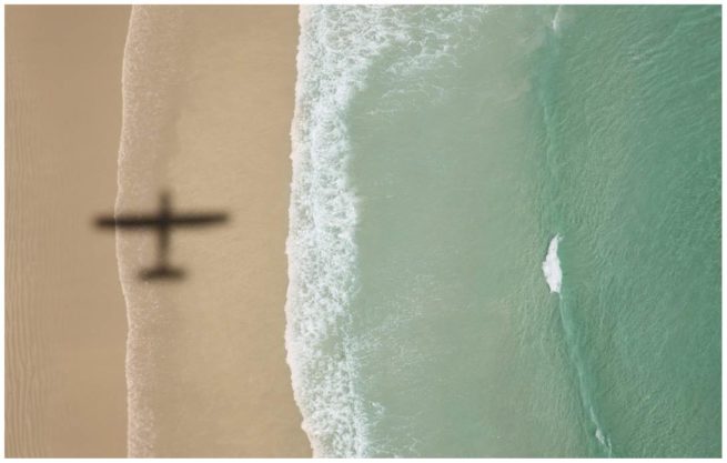 Luftaufnahme von einem Strand mit Brandung. Das Flugzeug wirft einen Schatten auf den Strand.