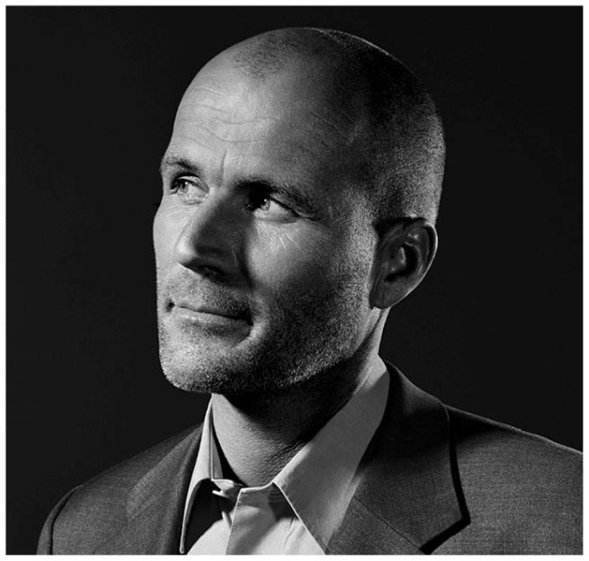 Schwarz weiss Studioportrait von einem Mann mit Glatze im Anzug vor schwarzem Hintergrund