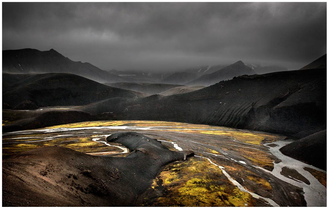 Landschaft auf Island mit schwarzen Hügeln und gelbem leuchtenden Bewuchs im Vordergrund bei bedrohlichem Wetter